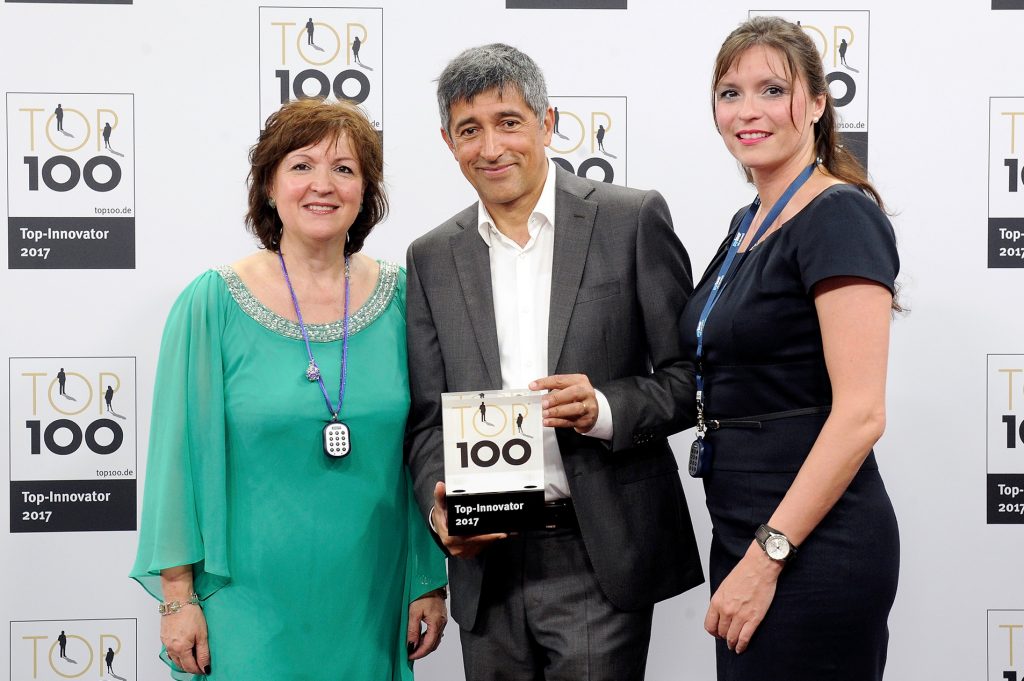 Zwei Frauen mit einem Mann, der einen Preis in den Händen hält, vor einer Werbewand mit der Aufschrift 100 Top-Innorator 2017.